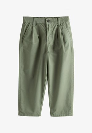 Junge mit blondem Haar steht mit einer Hand in der Tasche, trägt ein weißes T-Shirt, olivgrüne Hose und weiße Sneakers vor beigem Hintergrund.