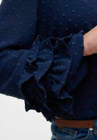 Blusa blu navy con tessuto testurizzato e piccoli motivi a pois, e maniche drammatiche a volant. Il materiale sembra leggero e fluido.