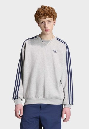 Mladý muž s kudrnatými červenými vlasy, oblečený v šedé mikině Adidas s tmavě modrými pruhy a tmavě modrých kalhotách, stojící před jednoduchým světlým pozadím.
