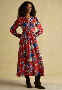 Rotes Blumenmusterkleid mit langen Ärmeln, V-Ausschnitt, geraffter Taille und bunten Mustern mit blauen und lila Blumen, kombiniert mit braunen Stiefeln.