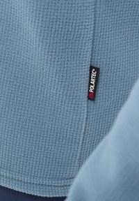 Pull en polaire bleu clair, avec une surface texturée et un ourlet cousu. Comprend une étiquette noire en Polartec®.