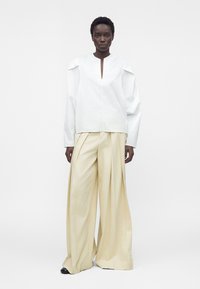 Mannequin portant un haut blanc à manches longues avec une fente au niveau de l'encolure devant et un pantalon plissé beige à jambes larges sur un fond uni.