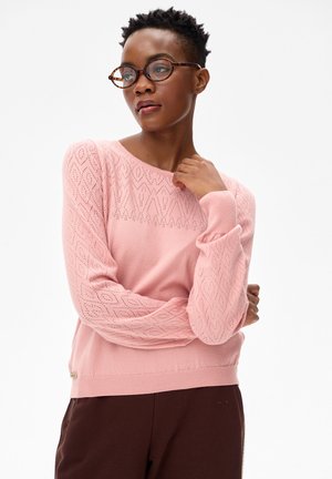 Kobieta o krótkich włosach, nosząca okulary w panterkę i różowy, teksturowany sweter, stoi ze skrzyżowanymi ramionami na białym tle.