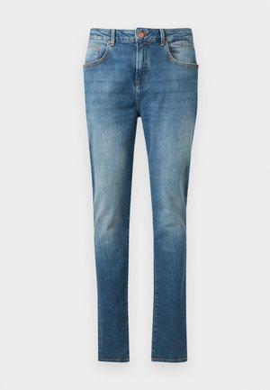 Sinised kitsalt lõikega teksapüksid esitaskute ja kerge pleekimisega reitel ja põlvedel, esitatud ühevärvilise heleda tausta ees.