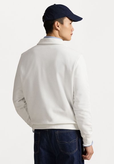 Valge tõmblukkidega jakk kraega, sile tekstuur ja pikad varrukad. Sobitatud tumedate siniste teksadega. Tumesinine müts on pandud tagurpidi.