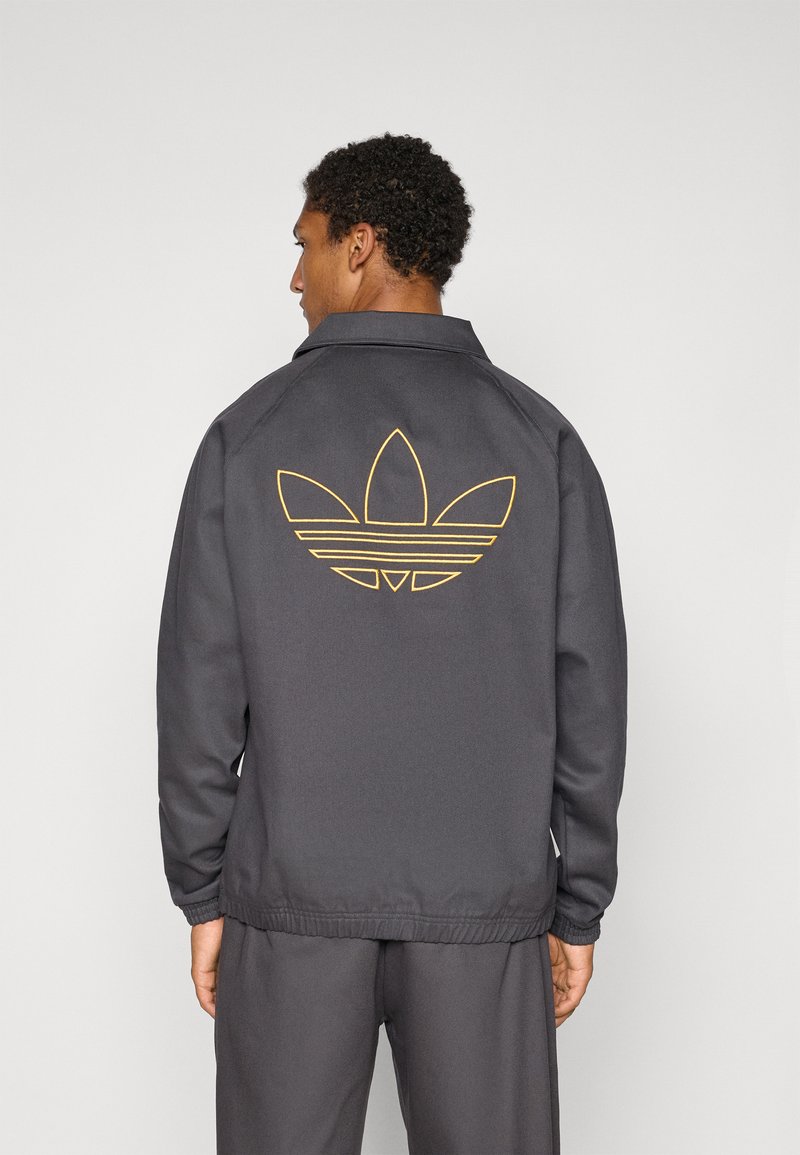 Juoda striukė su lygiu paviršiumi, ant nugaros pasižyminti dideliu auksiniu Adidas logotipu, su apykakle ir elastingais rankogaliais.