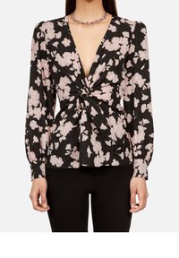 Zwarte blouse met lange mouwen en roze bloemenprint, diepe V-hals, gedraaide knoop aan de voorkant, gedragen met zwarte broek en een paarse kraal ketting.