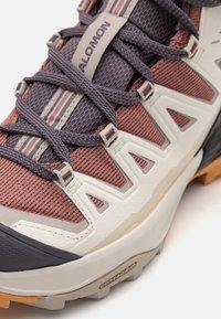 Trailrunningschuh mit einem hellgrauen und kastanienbraunen Mesh-Obermaterial, griffiger Gummisohle, lila Akzenten und einer verstärkten Zehenkappe.