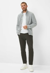 Grijze gebreide cardigan met een kraag, houten knopen, gedragen over een wit T-shirt, gecombineerd met donkergroene broek en witte sneakers.