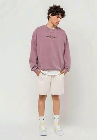 Joven con cabello rizado que lleva un sudadera malva, pantalones cortos beiges, calcetines blancos y zapatillas en tonos pastel, de pie con las manos en los bolsillos.
