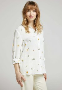 Chemise blanche à boutons, fabriquée en tissu texturé, ornée de broderies florales colorées en jaune et vert. Manches roulées et coupe décontractée.