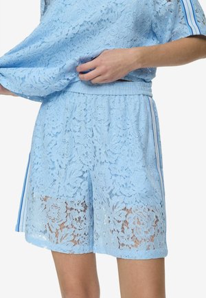 Pantalones cortos de encaje floral azul claro con cintura elástica y rayas laterales, usados por una persona que levanta ligeramente una blusa de encaje a juego.