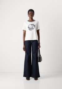 T-shirt bianco in cotone con grafica blu navy, abbinato a pantaloni larghi in denim scuro. Il modello tiene una piccola borsa nera, indossando sandali neri.