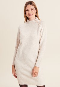 Robes en tricot crème, col montant côtelé, manches longues, avec des accents de boutons dorés sur une épaule, présentant un tissu texturé.