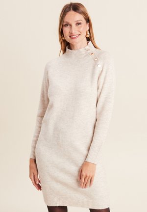 Brede gebreide sweatjurk in crèmekleur, met een geribbelde hoge hals, lange mouwen en gouden knoopaccenten op één schouder, vervaardigd uit een gestructureerde stof.