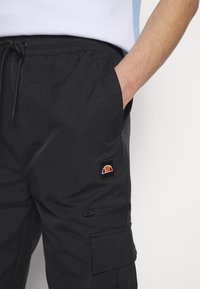Personne portant un pantalon cargo noir avec taille élastique et cordon, main dans la poche, montrant un écusson logo et un détail de poche zippée.