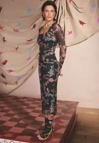 Vestito nero aderente con motivo floreale, maniche lunghe trasparenti; fiori e uccelli vivaci; abbinato a calzini neri e tacchi con cinturini gialli.