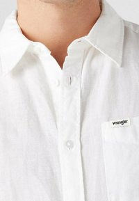 Biała lniana koszula z guzikami, z wyraźnym kołnierzykiem, kieszonką na przodzie i małą czarną metką "Wrangler" na kieszeni.