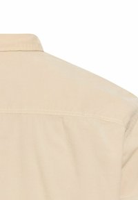 Beige overhemdbloes met een gestructureerde stof, achteryoke naad en eenvoudig kraagontwerp, met zichtbare stikdetails.
