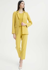 Żółta marynarka dopasowana z odpowiadającymi spodniami, wykonana z gładkiego materiału. Posiada klapy w kształcie notcha, kieszenie i lekko luźny krój.