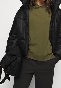 Osoba oblečená v olivově zelené mikině a černých kalhotách, držící černou prošívanou bundu přehozenou přes ruku na jednobarevném pozadí.