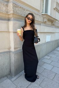 Svart, ribbad maxi-klänning med tunna axelband, kompletterad med en svart handväska och solglasögon; håller i en gul kaffekopp.