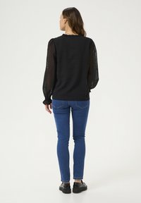Czarny top z długim rękawem, z przezroczystymi, kropkowanymi rękawami i falbaniastym dekoltem, połączony z niebieskimi skinny jeansami i czarnymi butami.