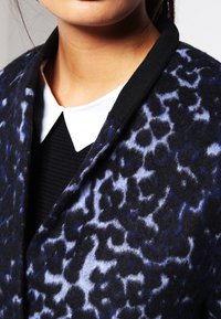 Donna che indossa un cappotto nero e blu a fantasia sopra una maglietta nera con un colletto bianco a punta, mostrando solo il collo e la parte inferiore del viso.