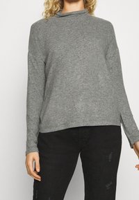 Szary top z długim rękawem i wysokim kołnierzem, wykonany z miękkiego materiału. Posiada luźny krój i boczne rozcięcia, zestawiony z czarnymi, zniszczonymi spodniami.