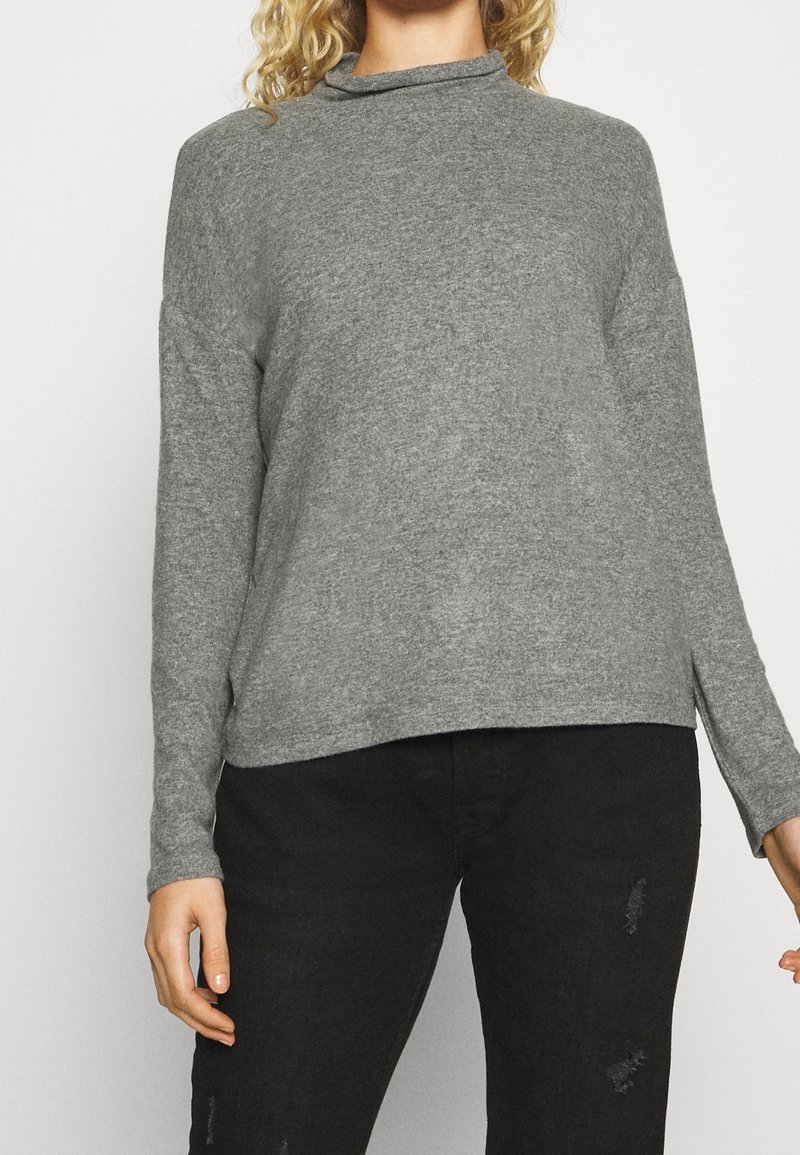 Szary top z długim rękawem i wysokim kołnierzem, wykonany z miękkiego materiału. Posiada luźny krój i boczne rozcięcia, zestawiony z czarnymi, zniszczonymi spodniami.