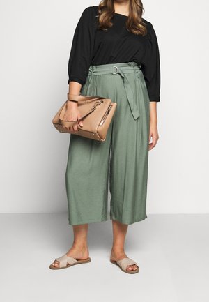 Femme portant un pantalon vert ample et court, un haut noir à manches trois-quarts, des sandales beiges, tenant un sac à main beige à côté d'elle.