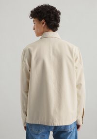 Beige katoenen jas met een relaxte pasvorm, gestructureerde kraag en lange mouwen. De achterkant heeft een eenvoudige uitstraling met subtiele stiksels.