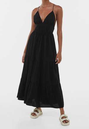 Femme portant une robe maxi noire sans manches avec fines bretelles et jupe à volants, associée à des sandales blanches ouvertes avec des boucles.