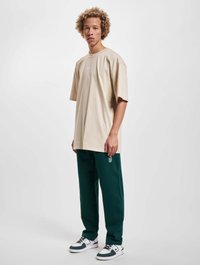 Beige oversized t-kreksa ar īsām piedurknēm, kombinēta ar tumši zaļiem treniņbiksēm ar logotipa detaļu un melnbaltiem sporta apaviem.