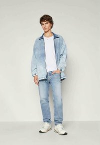 Jaqueta de denim azul claro com fecho de zip, t-shirt branca por baixo e jeans azul claro. Ténis brancos desportivos completam o visual.