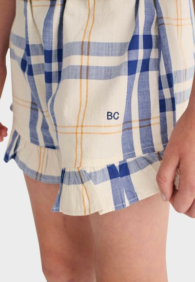Shorts à carreaux bleus, beiges et marron clair, avec des ourlets à volants et le monogramme brodé "BC" près de la cuisse droite, portés par une personne debout.