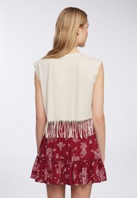 Top senza maniche color crema con dettagli in frange lungo l'orlo, abbinato a una gonna a più strati con motivo floreale rossa.