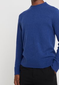 Niebieski sweter z wełny z okrągłym dekoltem, ściągaczami na mankietach i u dołu, gładką fakturą oraz dopasowanym krojem. Noszony na czarnych spodniach.