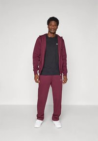 Borszínű kapucnis pulóver és pamutkeverék melegítőnadrág, fekete pólóval párosítva. Mellkason és nadrágon logóval díszítve. Fehér cipő.