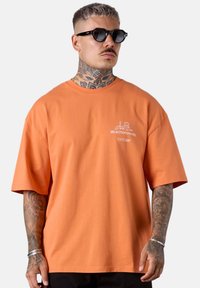 T-shirt en coton orange avec une coupe décontractée, dotée de manches courtes et d'un petit logo blanc sur la partie gauche de la poitrine. Texture unie.