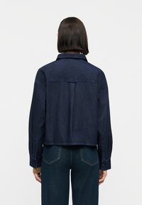 Άτομο με μεσαίου μήκους σκούρα μαλλιά, φορώντας σκούρο μπλε κοντό τζιν μπουφάν και μπλε τζιν, με την πλάτη προς το μέρος μας και λευκό φόντο.