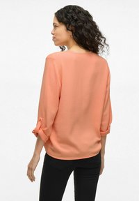 Blusa leggera color corallo con maniche a tre quarti e polsini arrotolati; tessuto testurizzato con vestibilità comoda, abbinata a pantaloni neri.