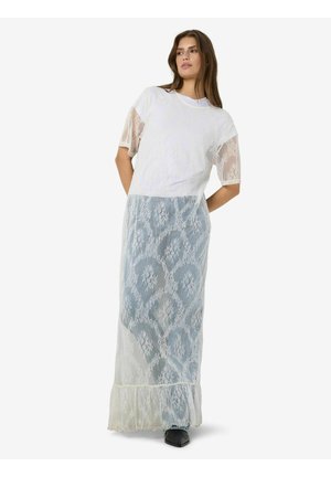 Femme portant un haut court en dentelle blanche et une longue jupe en dentelle par-dessus un pantalon bleu avec des chaussures noires pointues, debout les mains dans le dos.