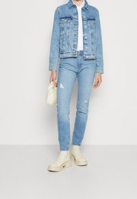 Chaqueta de mezclilla azul claro con dos bolsillos en el pecho y cierre de botones, combinada con jeans azul claro desgastados y botines crema. Sosteniendo un bolso blanco.