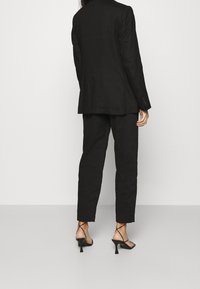 Blazer noir ajusté et pantalon assorti en tissu léger. Portés avec des sandales à talons fines. Vue arrière mettant en valeur les lignes épurées et la coupe.