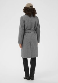 Manteau en laine grise avec une taille ceinturée, des manches longues et une silhouette droite ; assorti à un pantalon noir et des bottes à talons.
