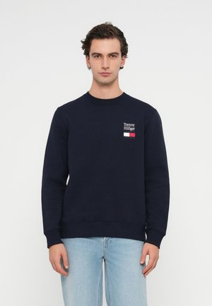 Mężczyzna ubrany w granatową bluzę Tommy Hilfiger i jasnoniebieskie dżinsy, stojący na jednolitym białym tle.