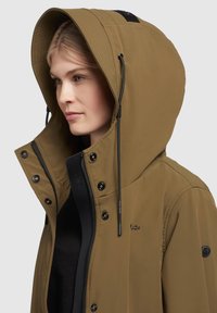 Veste imperméable marron avec une grande capuche ajustable, quincaillerie noire, fermeture éclair et boutons, présentant une silhouette épurée et élancée.