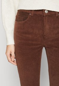 Rękaw kremowego swetra i dłoń obok brązowych spodni z kordurowej tkaniny z przednimi kieszeniami i srebrnym zapięciem.