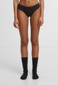 Fekete pamut bikini alsónemű, alacsony derék vonalú kialakítással, sima textúrával és elasztikus derékrésszel. A modellen azonos színű fekete, lógós zoknik találhatók.